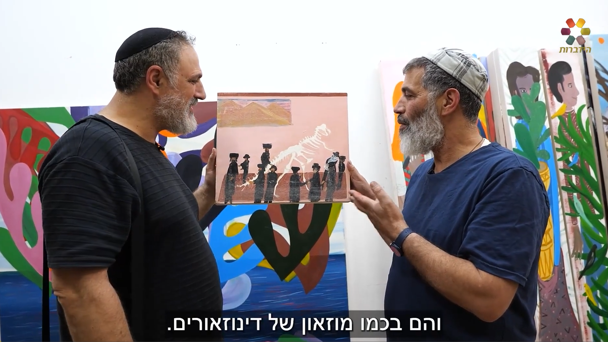 Two bearded men wearing kippahs (one in a black shirt and the artist Shai Azoulay in a blue shirt) discuss a small painting featuring Haredi figures next to a dinosaur skeleton, set against a backdrop of large, colorful canvases.
