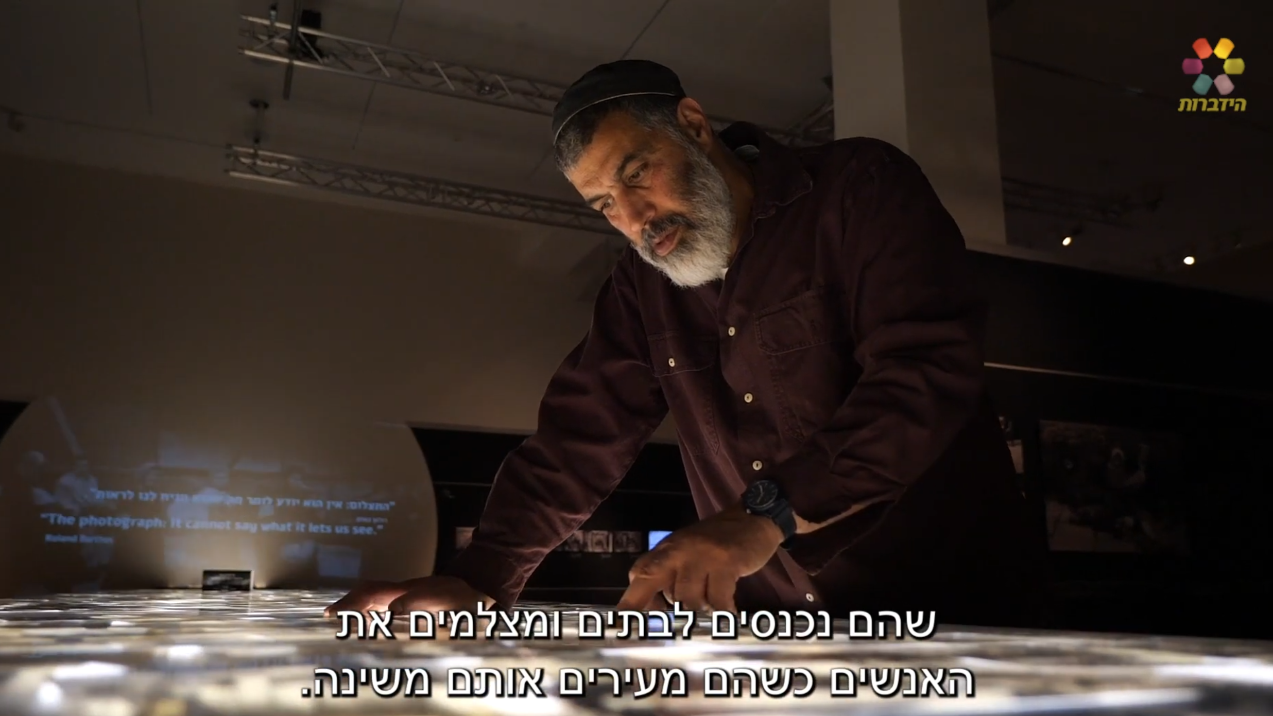 A man reviewing photographs on a light table at an exhibition, from a Hidabroot channel broadcast